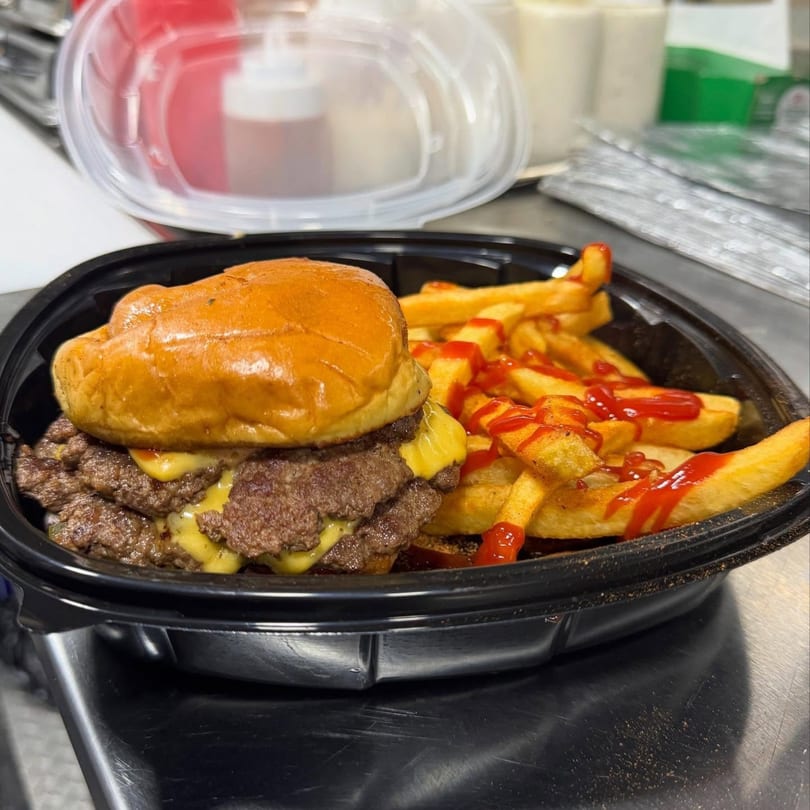 A double cheeseburger with melted cheese and a side of ketchup-drizzled fries in a black takeout container.