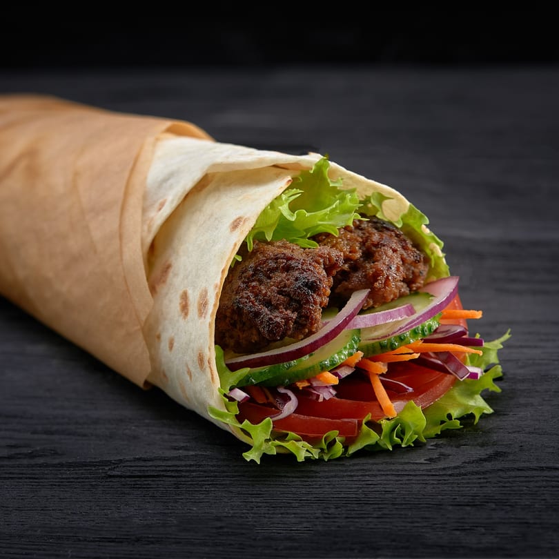 Halal beef kofta wrap with fresh lettuce, tomatoes, onions, cucumbers, and carrots on a dark surface.